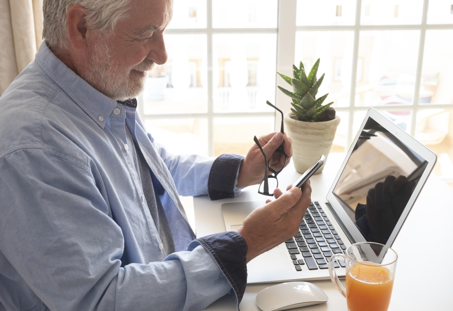 Bearded senior man stays at home using cellphone. Desk office with laptop computer and orange juice glass.Seniors and technology concept. Intense light from the window